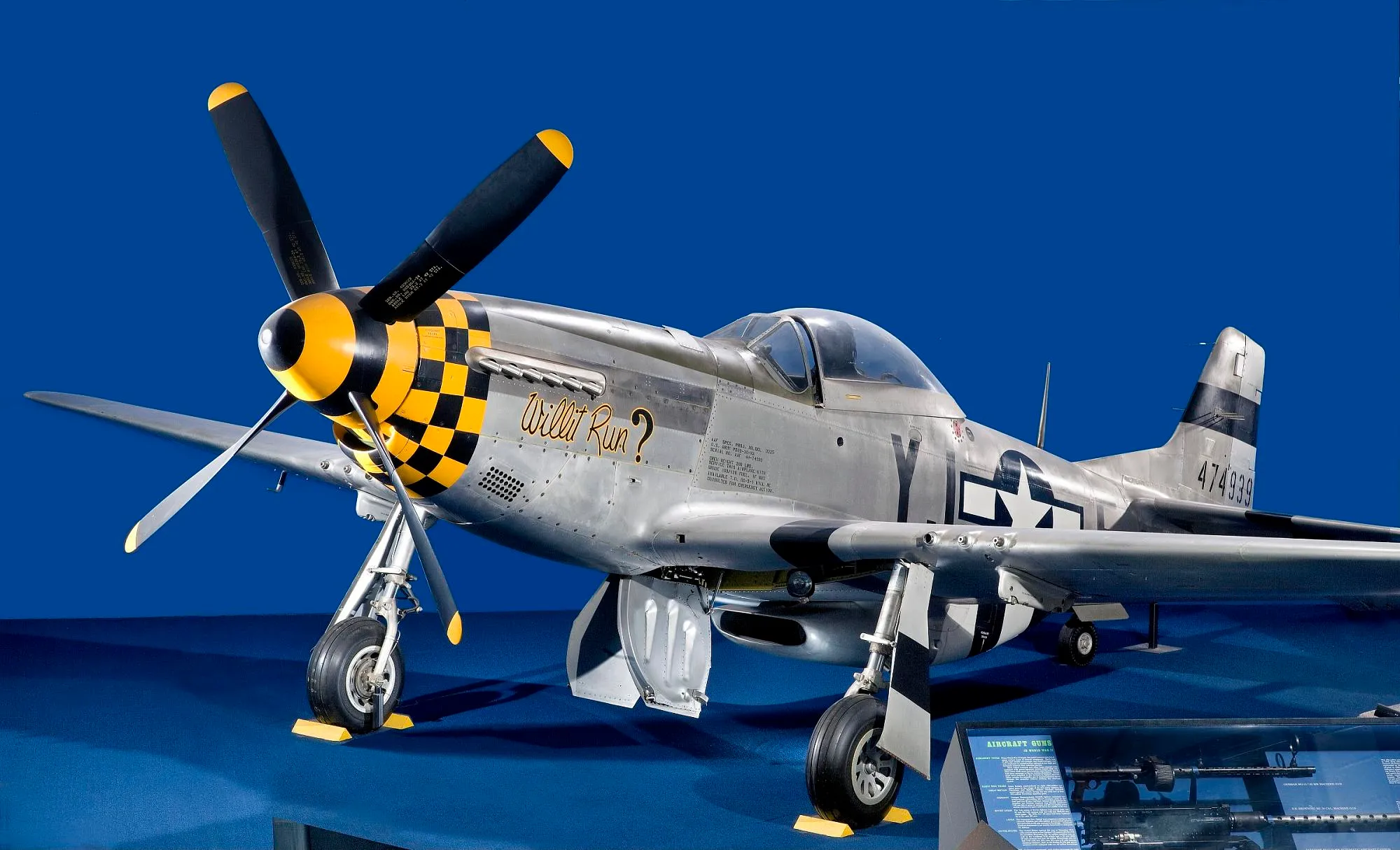A silver North American P-51D Mustang with a yellow and black checkered nose and the name Willit Run? displayed on a blue background at the National Air and Space Museum